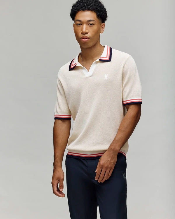 A man wears the Psycho Bunny MENS CARTER JOHNNY COLLAR KNIT POLO (MT0400093), a cream 100% cotton short-sleeve polo with red and black striped Johnny collar and sleeve trim, paired with navy pants, against a light gray background.