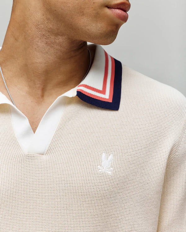 A close-up shows a person wearing the Psycho Bunny MENS CARTER JOHNNY COLLAR KNIT POLO (MT0400093), a cream 100% cotton knit polo with blue, red, and white striped Johnny collar and an embroidered white logo on the chest. Only the lower face and neck appear.