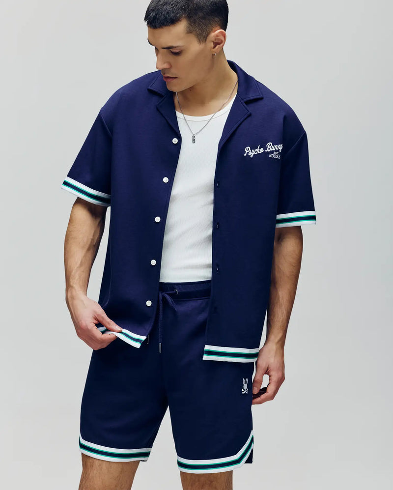 A man wears the Psycho Bunny MENS BASIL TAPED CAMP COLLAR SHIRT - MT0500004 in navy, featuring a Johnny collar and white-teal trim over a white tank, paired with matching shorts. He looks down, holding the hem of his premium cotton-modal blend shirt.
