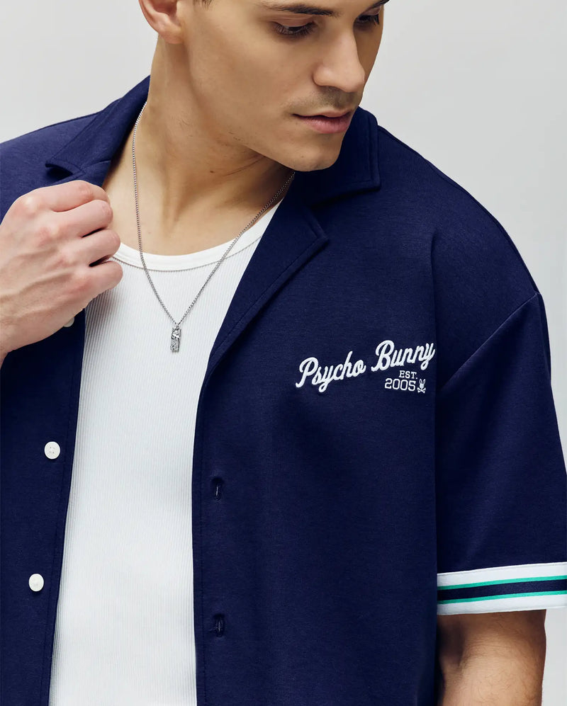 A man wears the Psycho Bunny MENS BASIL TAPED CAMP COLLAR SHIRT (MT0500004) in navy blue, made from a cotton-modal blend with “Psycho Bunny Est. 2005” embroidery, over a white tank and silver necklace, looking down to his left.