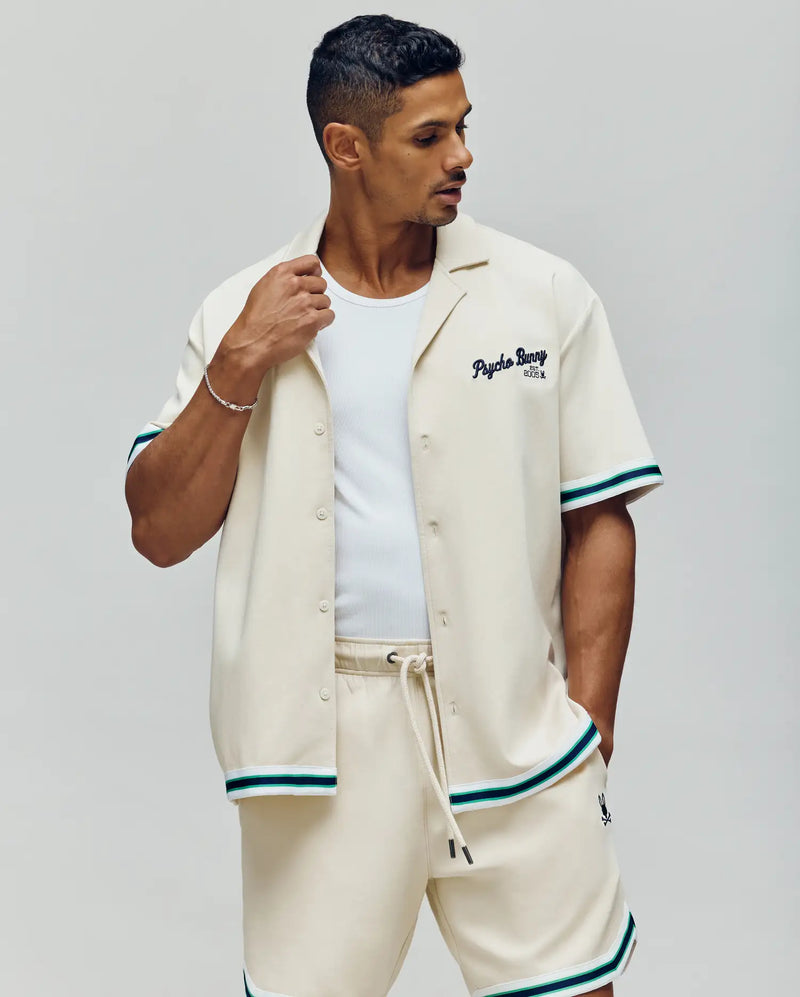 A man wears the Psycho Bunny MENS BASIL TAPED CAMP COLLAR SHIRT (MT0500004) in a cream cotton-modal blend with green and black striped trim and Johnny collar, paired with matching shorts and a white tank, standing against a plain light background.