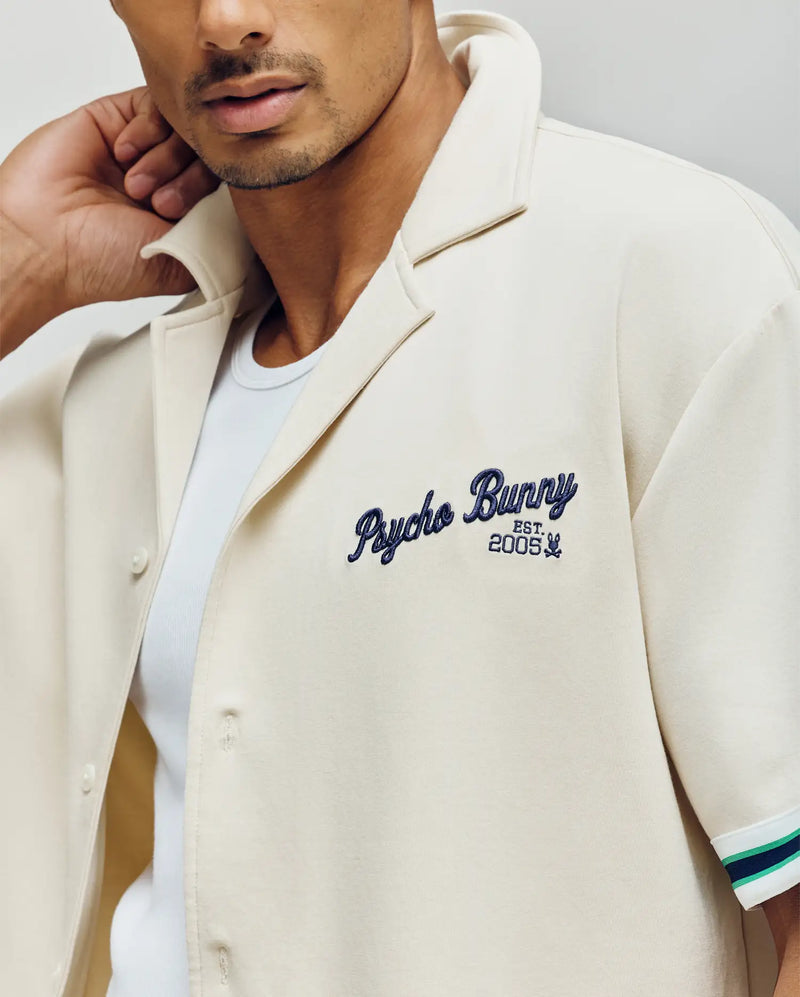 A man wears the Psycho Bunny MENS BASIL TAPED CAMP COLLAR SHIRT (MT0500004) in light cream, made from a premium cotton-modal blend. The shirt features a Johnny collar and blue "Psycho Bunny EST. 2005" chest embroidery.