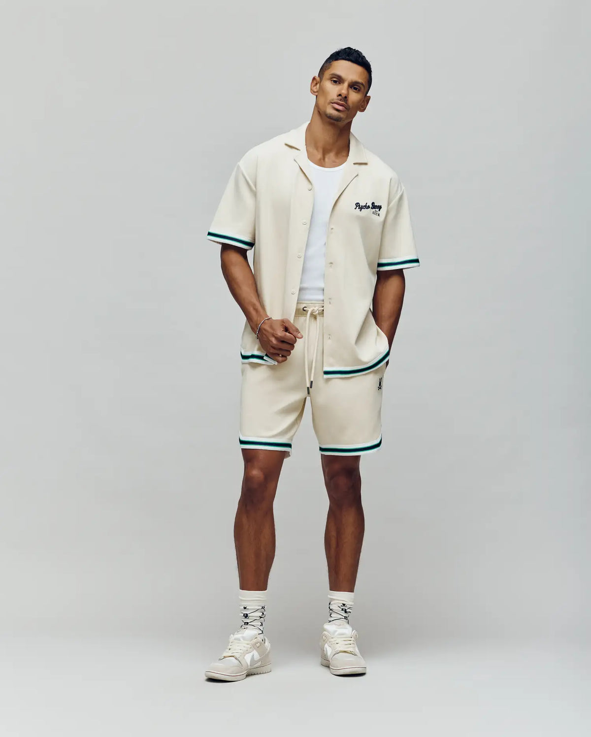 A man stands confidently in the Psycho Bunny MENS BASIL TAPED CAMP COLLAR SHIRT (MT0500004), paired with matching cream shorts with green trim, a white tank, cream sneakers, and patterned white socks against a neutral background.