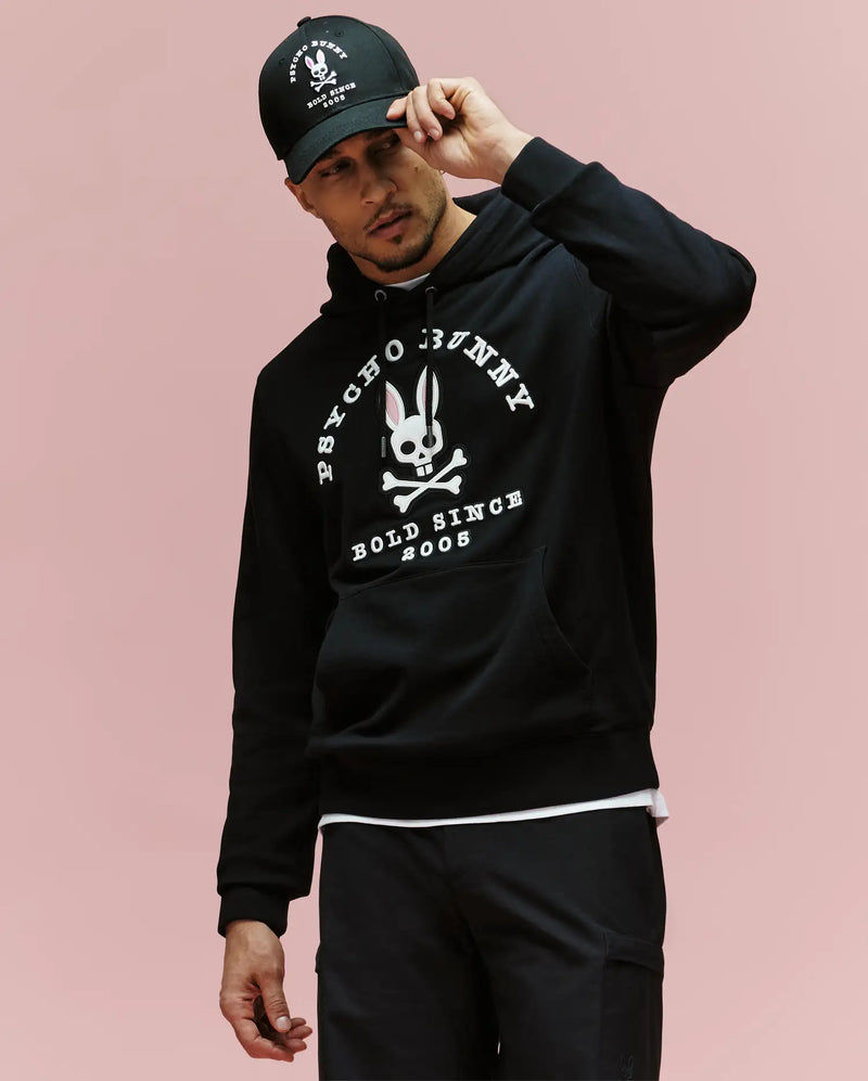 A man in a black Psycho Bunny MENS ARCHIVE HOODIE - MT0500009 and matching cap, both with white skull-and-bunny logos and "Psycho Bunny Bold Since 2005" text, stands against a light pink background.
