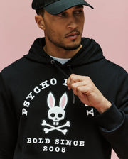 A man wears the Psycho Bunny MENS ARCHIVE HOODIE - MT0500009 in black, set against a pink background. Made from 100% cotton French terry, it features a bunny skull-and-crossbones faux-leather applique and "PSYCHO" & "BOLD SINCE 2005" text.