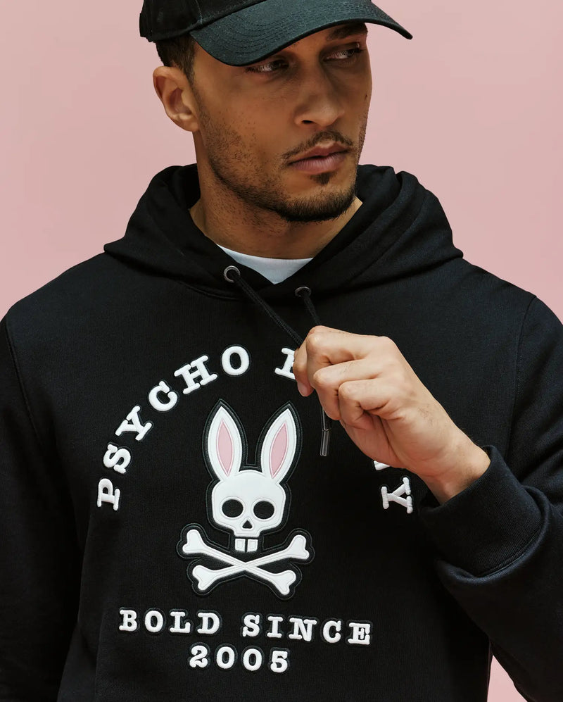 A man wears the Psycho Bunny MENS ARCHIVE HOODIE - MT0500009 in black, set against a pink background. Made from 100% cotton French terry, it features a bunny skull-and-crossbones faux-leather applique and "PSYCHO" & "BOLD SINCE 2005" text.