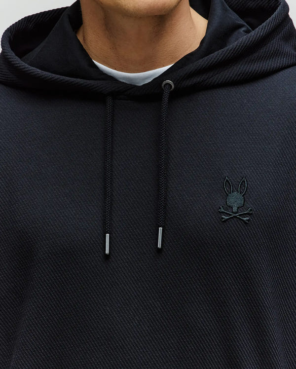 A close-up of a person in the Psycho Bunny MENS KAI HOODIE - MT0500019, black with twill texture, drawstring hood, and small embroidered Bunny logo on the left chest. A white shirt is worn underneath.