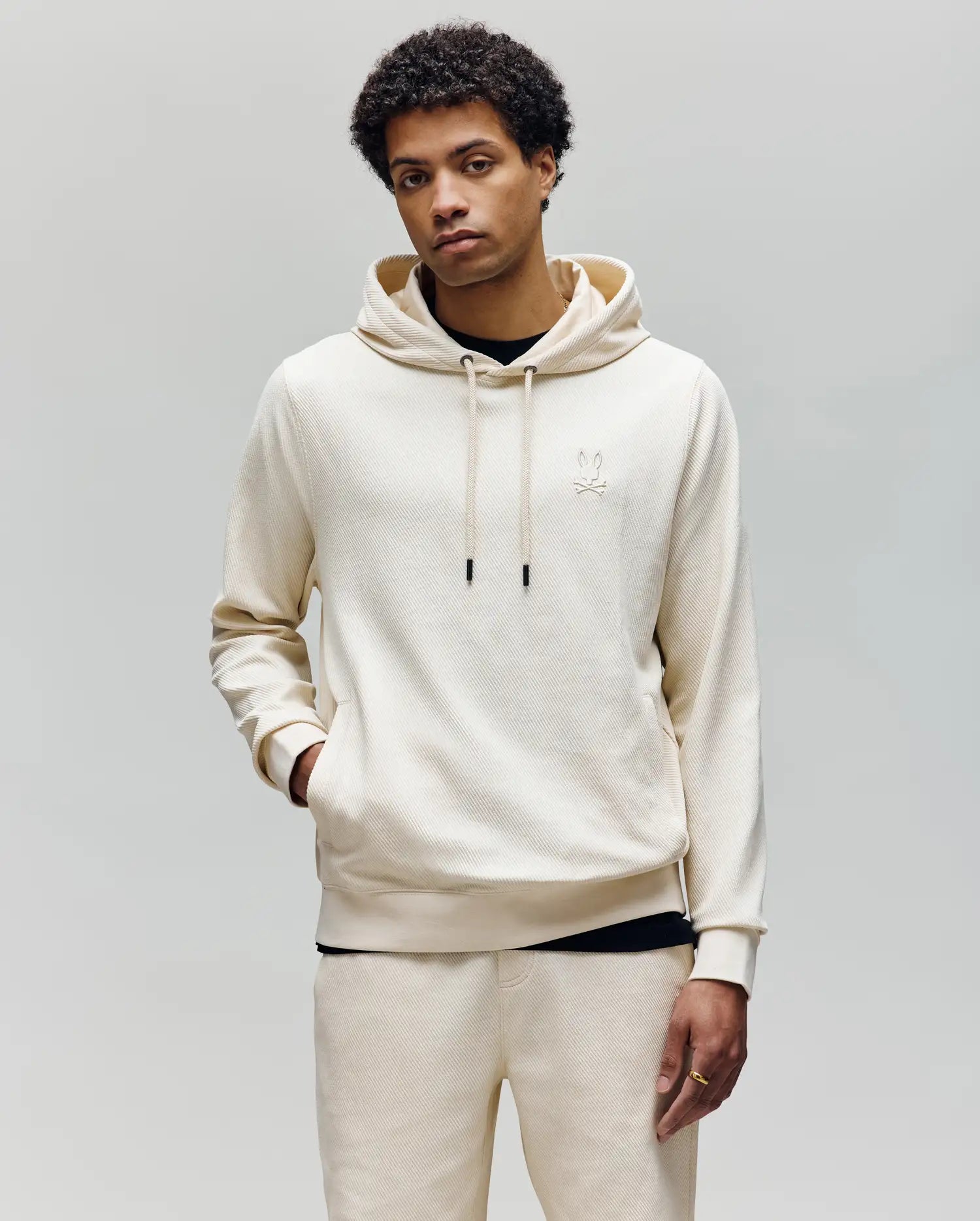 A young man with short curly hair wears the Psycho Bunny MENS KAI TWILL TEXTURE HOODIE (MT0500019) in cream, featuring an embroidered Bunny logo and matching sweatpants, standing against a plain light gray background.