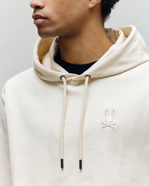 A person wears the Psycho Bunny MENS KAI TWILL TEXTURE HOODIE (MT0500019) in cream, featuring an embroidered Bunny logo and twill-textured fabric. The image shows only their lower face, neck, and upper torso.