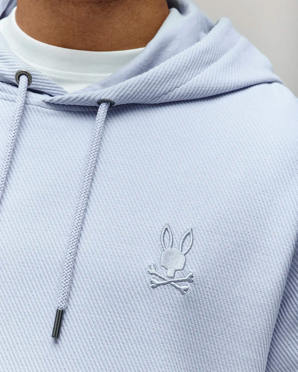 Close-up of a person wearing the Psycho Bunny MENS KAI HOODIE - MT0500019 in light purple with twill texture and a small embroidered Bunny logo on the chest, layered over a white shirt.