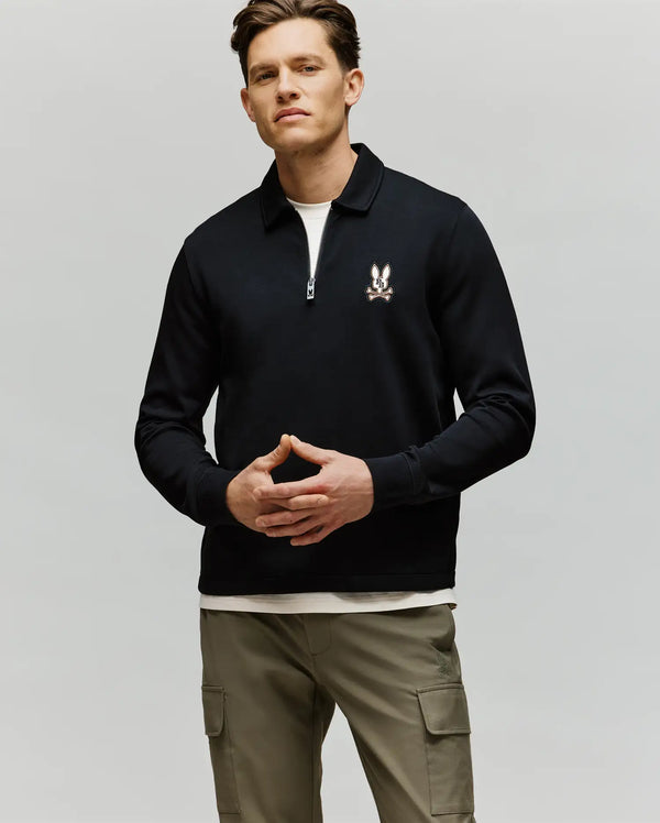 A man faces forward in the Psycho Bunny MENS DANIEL QUARTER ZIP SWEATSHIRT (MT0500023), a black pullover with a small chest logo, paired with a white undershirt and light khaki cargo pants, set against a plain light background.