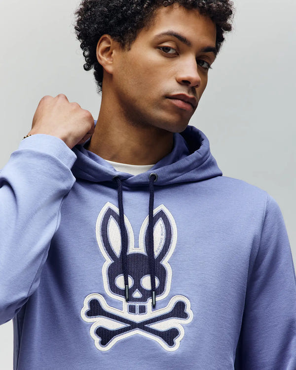 A person in the Psycho Bunny MENS RYDER EMBROIDERED BUNNY PATCH HOODIE (MT0500029) with a bold skull-and-crossbones bunny logo on the chest stands against a plain background, showcasing comfortable, laid-back style.