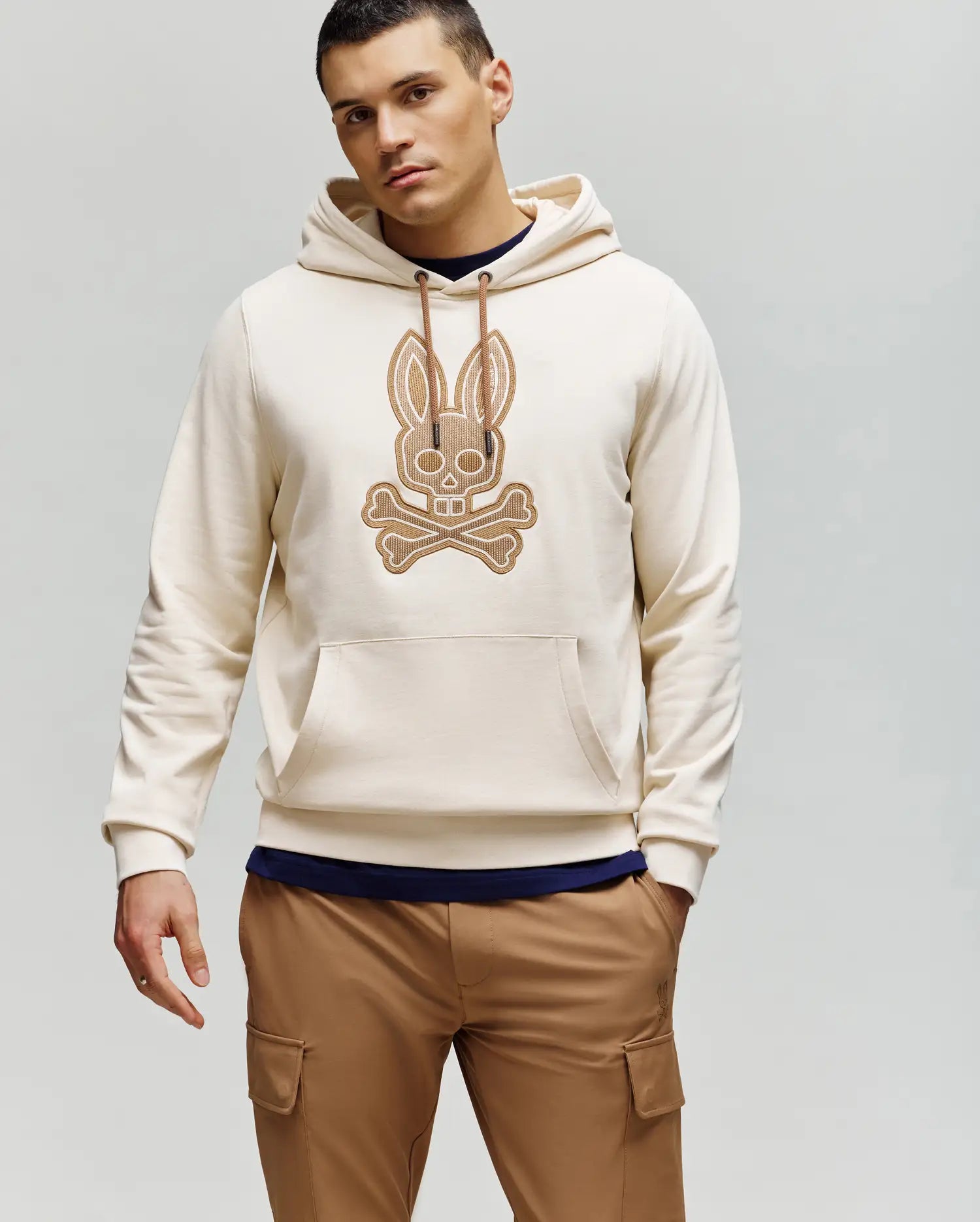 A man wears the Psycho Bunny MENS RYDER EMBROIDERED BUNNY PATCH HOODIE (MT0500029) in light beige, featuring an embroidered Bunny patch and crossbones on the front, styled with tan cargo pants against a plain light gray background.