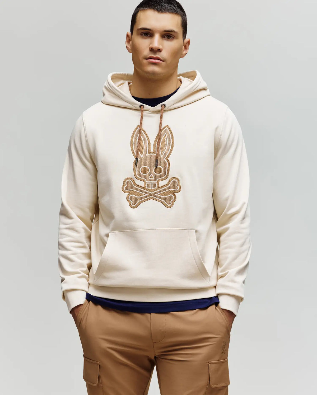 A man with short dark hair stands against a plain light background, wearing the Psycho Bunny MENS RYDER EMBROIDERED BUNNY PATCH HOODIE (MT0500029) in beige with a large crossbones graphic, paired with brown cargo loungewear pants.