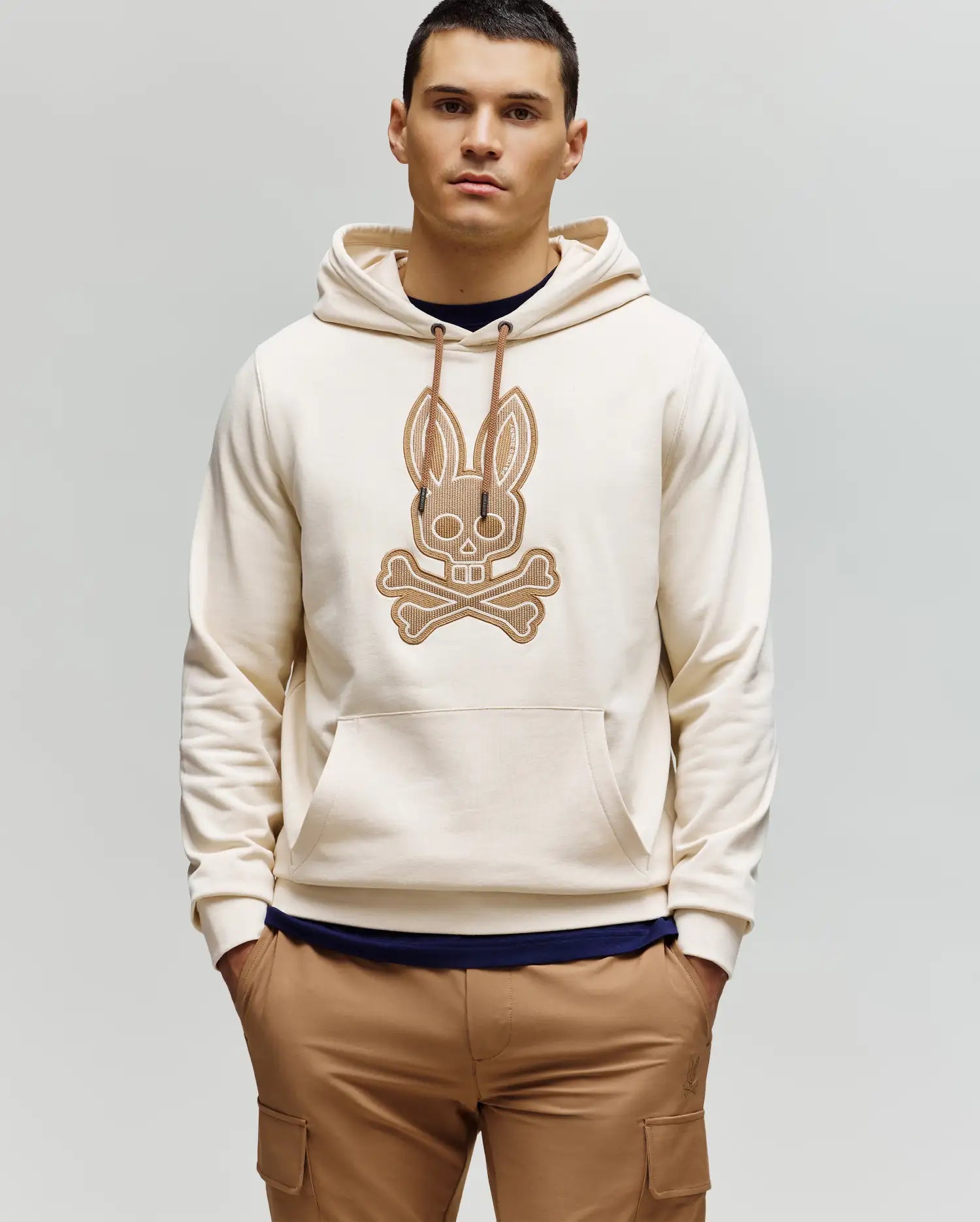 A man with short dark hair stands against a plain light background, wearing the Psycho Bunny MENS RYDER EMBROIDERED BUNNY PATCH HOODIE (MT0500029) in beige with a large crossbones graphic, paired with brown cargo loungewear pants.
