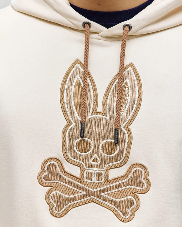 Close-up of Psycho Bunny’s cream-colored MENS RYDER EMBROIDERED BUNNY PATCH HOODIE (MT0500029), ideal for loungewear, with a large embroidered stylized bunny skull and crossbones patch outlined in brown thread.