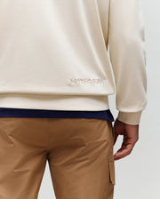 A person is seen from the back, waist down, wearing tan cargo pants and a cream Psycho Bunny MENS RYDER EMBROIDERED BUNNY PATCH HOODIE (MT0500029) featuring an embroidered Bunny patch near the hem for a stylish loungewear vibe.