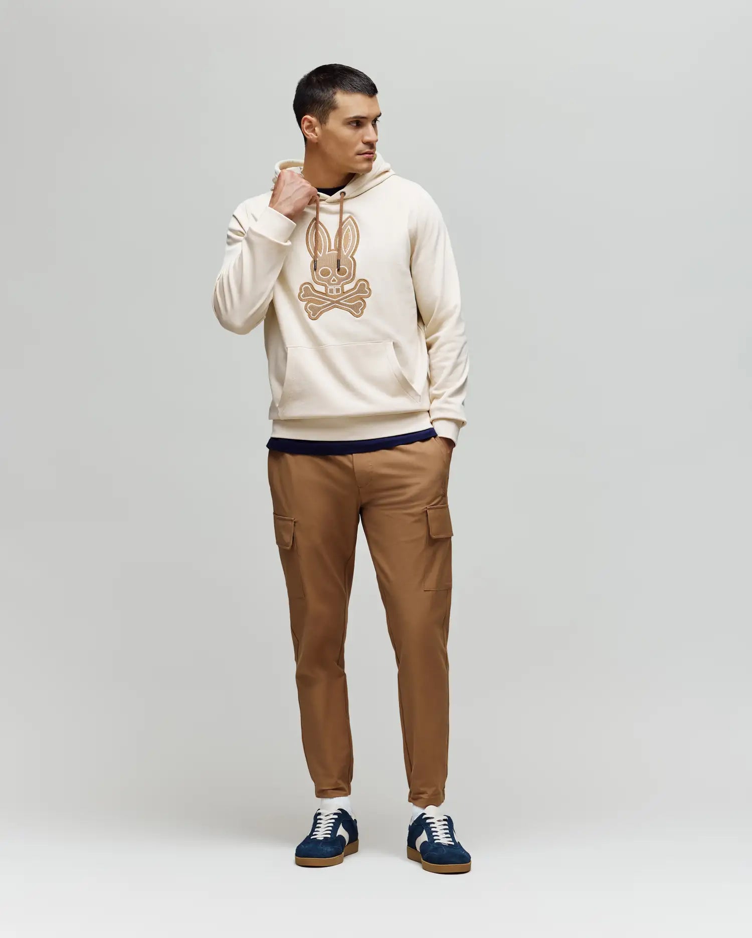 A man stands against a plain background wearing the Psycho Bunny MENS RYDER EMBROIDERED BUNNY PATCH HOODIE (MT0500029), paired with tan cargo pants and navy blue sneakers—ideal for a relaxed, laid-back look.
