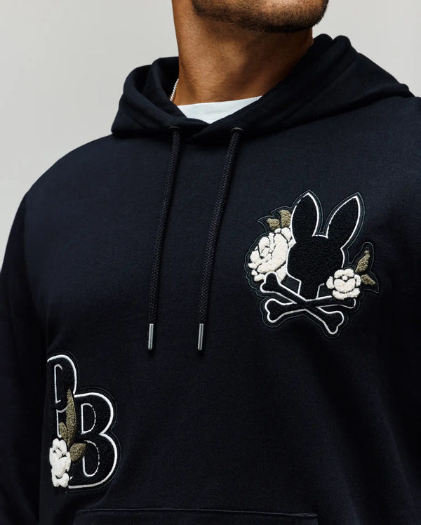 A person wears the Psycho Bunny MENS AUGUSTE HOODIE (MT0500043), a regular fit premium cotton hoodie with embroidered bunny head, crossbones, "PB" initials, chenille floral details, and partially out of frame face.