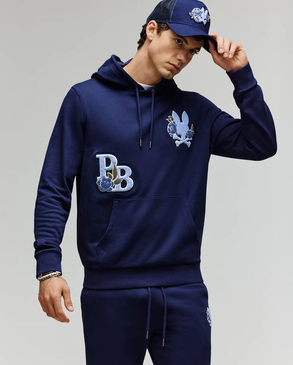 A man in a Psycho Bunny MENS AUGUSTE HOODIE (MT0500043), premium navy blue cotton with chenille floral details, matching sweatpants, and a navy cap stands against a plain light background, touching his cap's brim.