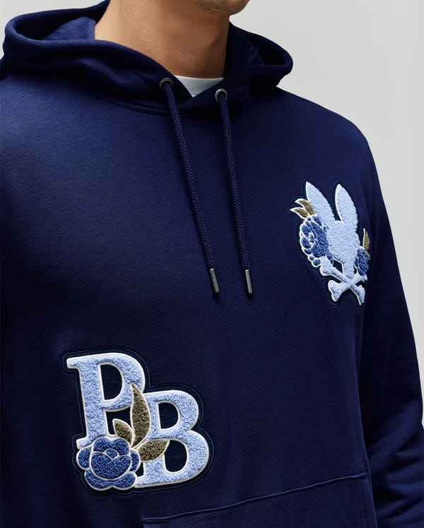 A person wears the Psycho Bunny MENS AUGUSTE HOODIE (MT0500043), a regular fit, navy premium cotton hoodie featuring embroidered rabbit and floral chest patches with "PB" letters, blue rose, and chenille floral accents.