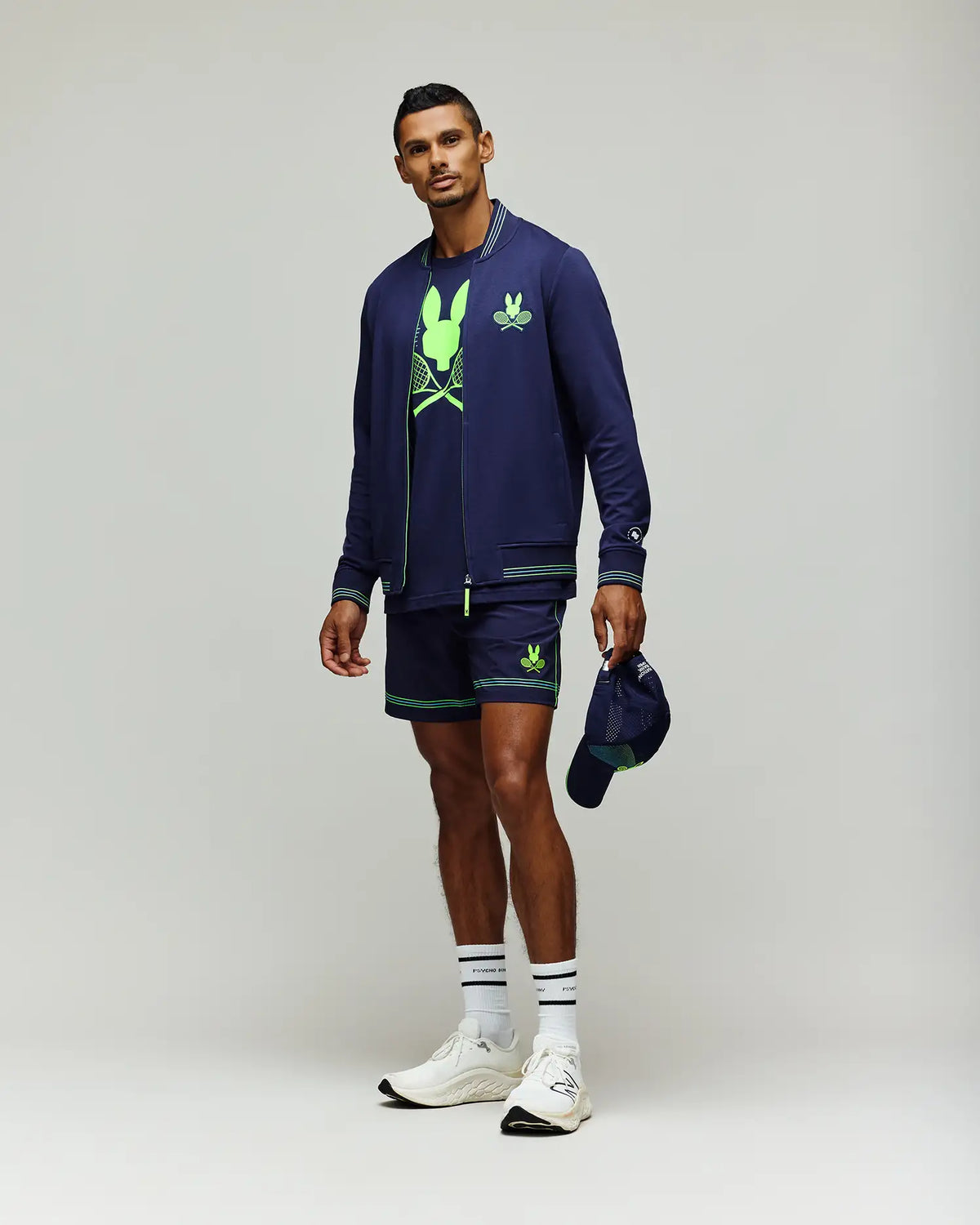 A man poses against a plain background wearing the Psycho Bunny Tennis Ball Crew Mens Jacket (MT0500049) in navy, matching shorts, a neon green graphic tee, white socks and sneakers, holding a navy cap in his left hand.