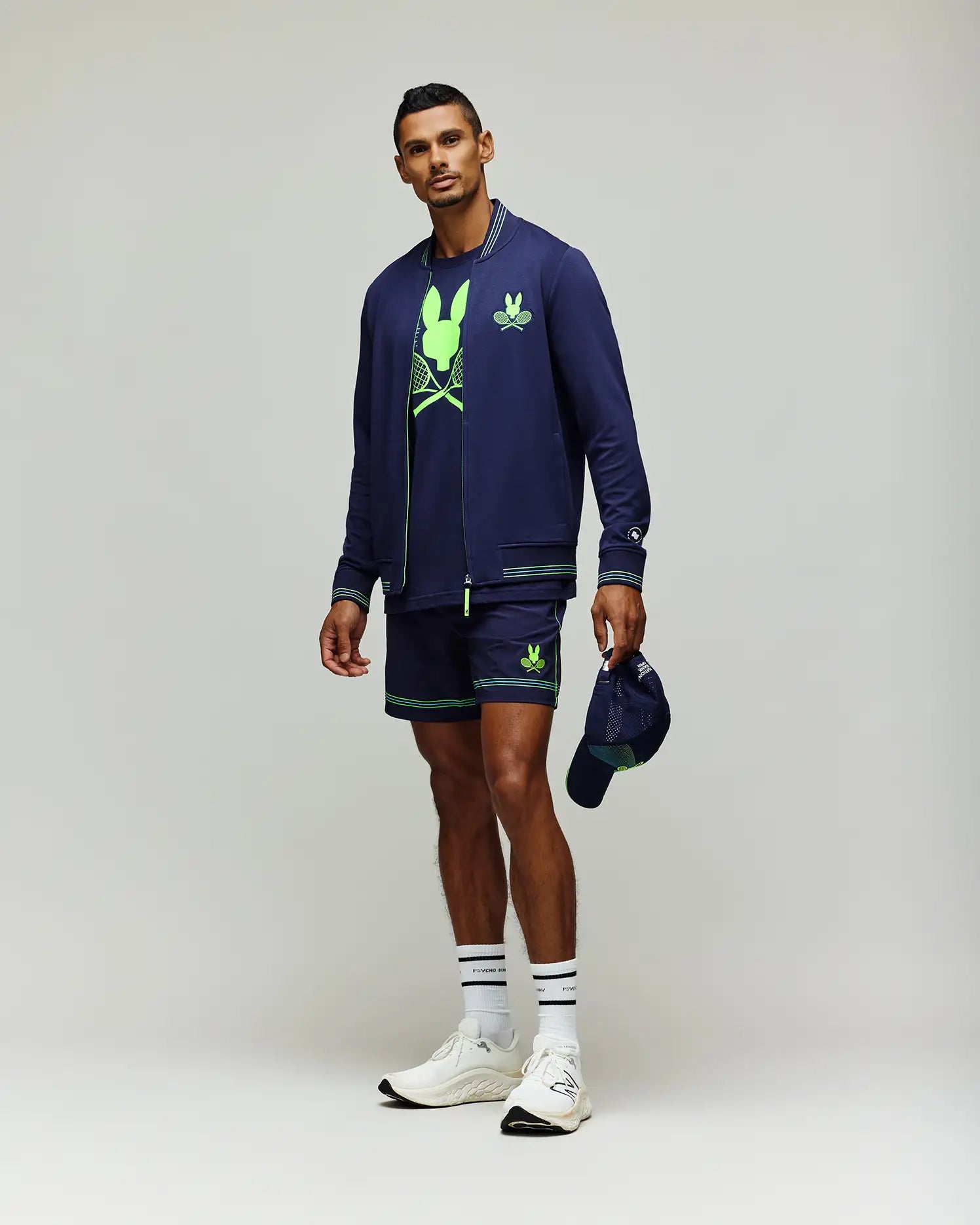 A man poses against a plain background wearing the Psycho Bunny Tennis Ball Crew Mens Jacket (MT0500049) in navy, matching shorts, a neon green graphic tee, white socks and sneakers, holding a navy cap in his left hand.