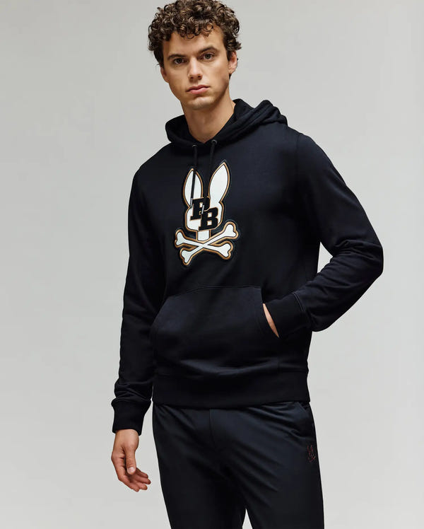 A young man wears the Psycho Bunny MENS WADE CORD PATCH HOODIE (MT0500112), an athletic hoodie in a premium cotton blend with an embroidered Bunny skull and crossbones logo, standing with one hand in his pocket against a plain background.