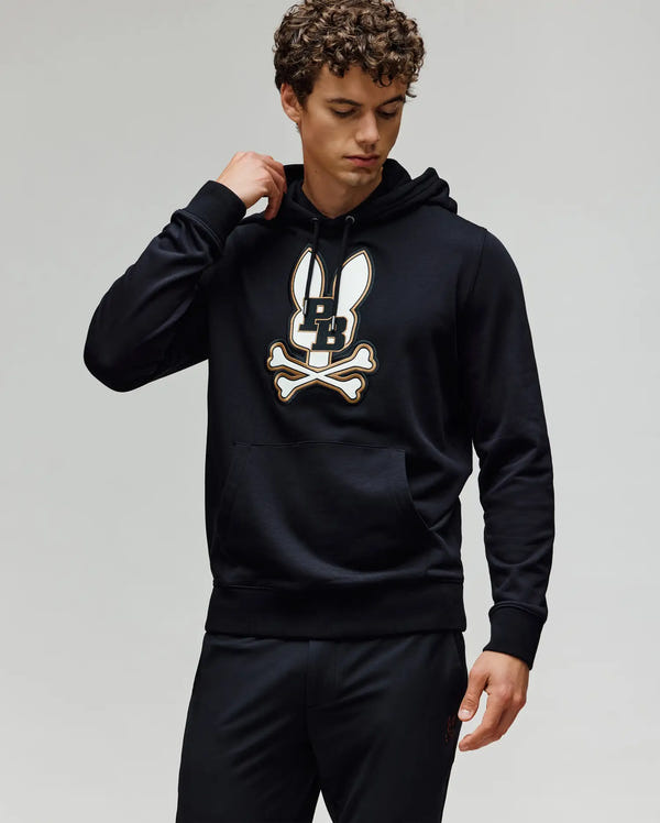A man with curly hair wears the Psycho Bunny MENS WADE CORD PATCH HOODIE (MT0500112) with an embroidered Bunny patch, paired with black pants, standing against a plain light background.