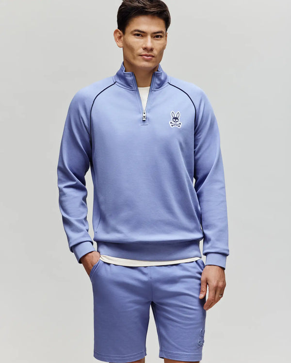 A man with short dark hair and a neutral expression stands against a light gray background wearing the Psycho Bunny MENS SHAWN 1/4 ZIP SWEATSHIRT (MT0500115) and matching shorts, both featuring the brand's cartoon bunny logo on the chest.