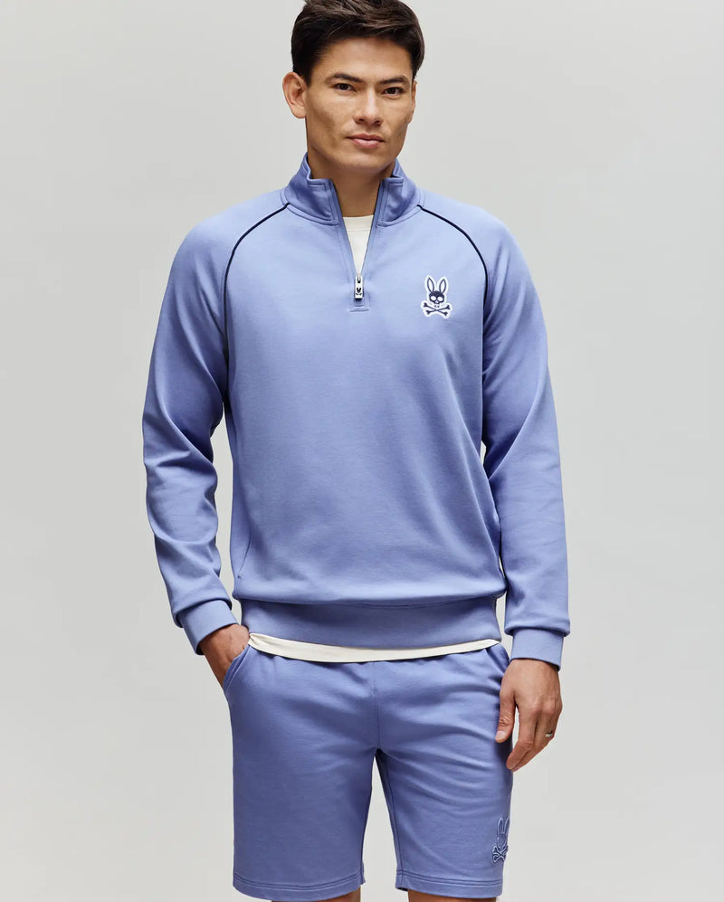 A man with short dark hair and a neutral expression stands against a light gray background wearing the Psycho Bunny MENS SHAWN 1/4 ZIP SWEATSHIRT (MT0500115) and matching shorts, both featuring the brand's cartoon bunny logo on the chest.