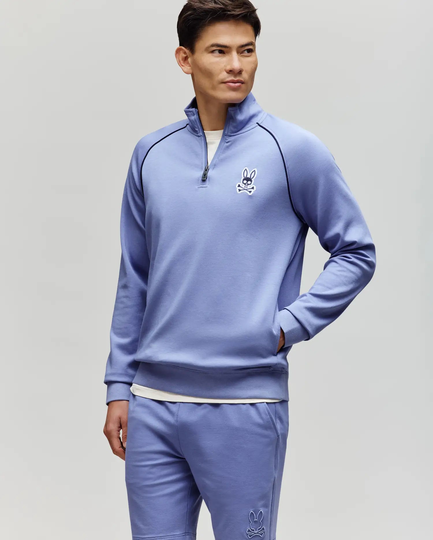 A man wears the Psycho Bunny MENS SHAWN 1/4 ZIP SWEATSHIRT (MT0500115) and matching shorts, both light purple with a white bunny logo, standing with one hand in his pocket against a plain light gray background.