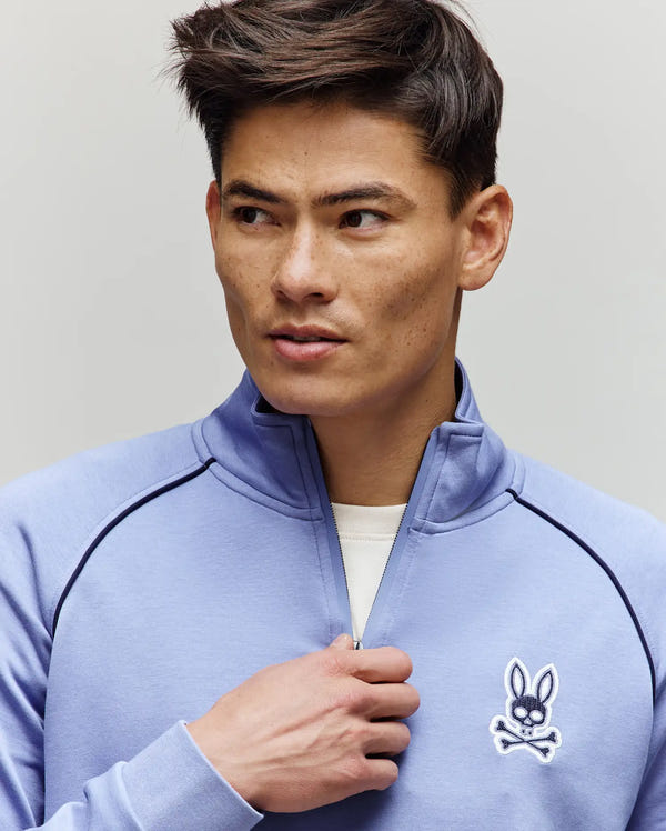 A man with short brown hair wears the Psycho Bunny MENS SHAWN 1/4 ZIP SWEATSHIRT (MT0500115), a light blue cotton-modal top with a bunny and crossbones logo on the chest. He holds the zipper, looking left against a plain, light background.