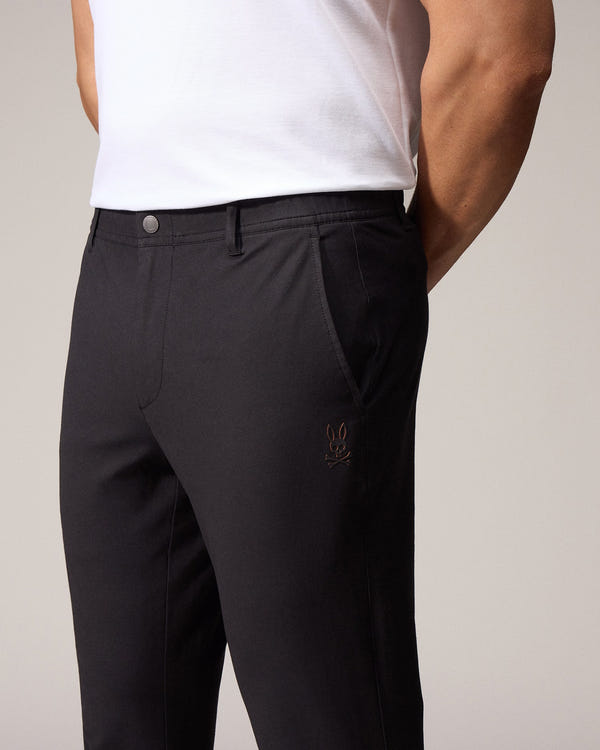 A close-up of a man in a light background wearing dark, tailored Psycho Bunny MENS MADISON COMMUTER PANTS with embroidered Bunny logos, focusing on the fit and muscular build of his lower body and biceps.