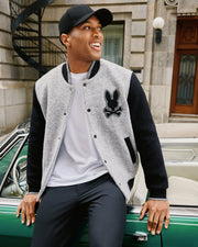 A person wearing a black cap, Psycho Bunny Men's Tom Wool Bomber Jacket (B6J744D200) in gray and black with a chenille embroidered bunny patch, white shirt, and black pants smiles while sitting on the edge of a green vintage car. The background includes a stone building with a spiral staircase and a wooden door.