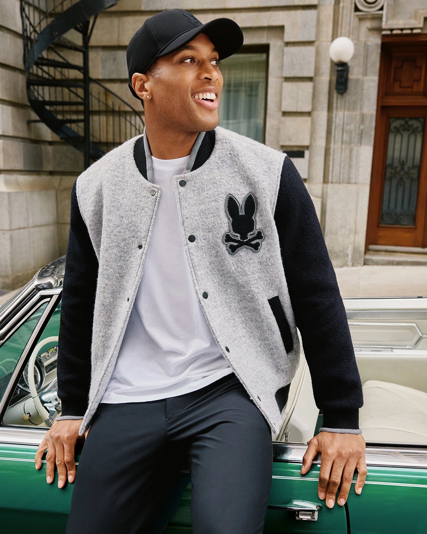 A person wearing a black cap, Psycho Bunny Men's Tom Wool Bomber Jacket (B6J744D200) in gray and black with a chenille embroidered bunny patch, white shirt, and black pants smiles while sitting on the edge of a green vintage car. The background includes a stone building with a spiral staircase and a wooden door.