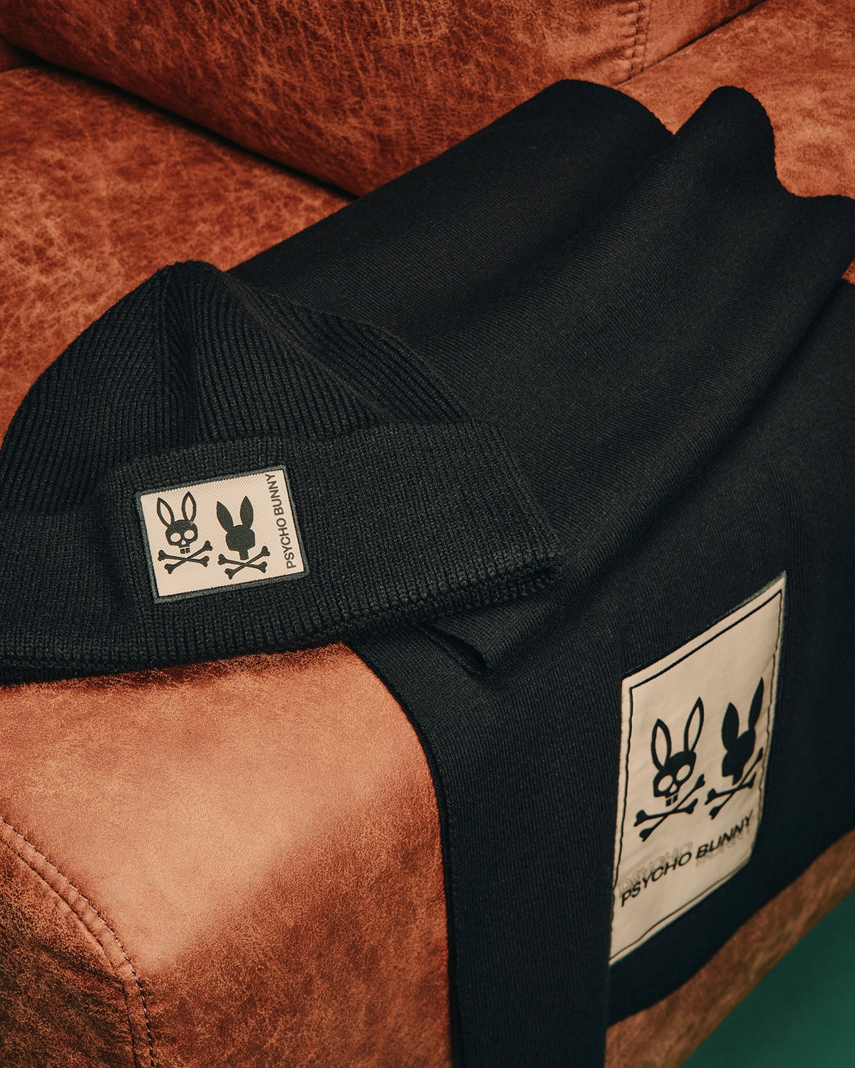 A black cotton-wool blend beanie and luxury Mens Monroe Twill Patch Scarf from Psycho Bunny, featuring white embroidered logos of a stylized bunny and skull crossbones, laid on a brown leather surface.