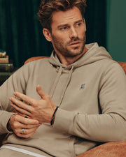 A man with short hair and a beard is sitting thoughtfully on a brown chair, wearing a Psycho Bunny MENS LEVY ESSENTIAL FLEECE HOODIE - B6H859D200. Rings adorn his fingers, while a green curtain and books create an intriguing backdrop.