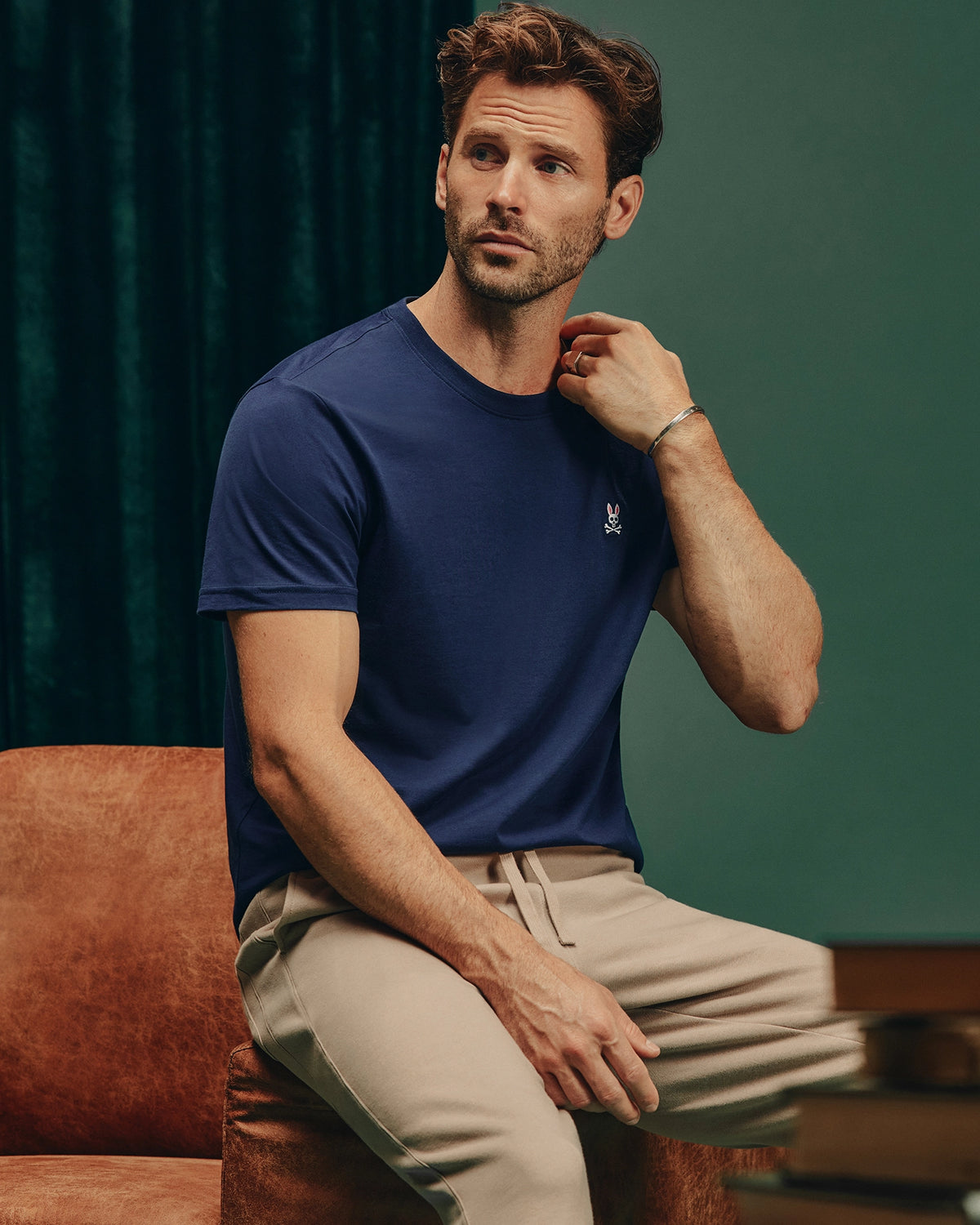 A man sits on a brown chair, wearing a versatile dark blue Psycho Bunny MENS CLASSIC CREW NECK TEE - B6U014CRPC made of 100% pima cotton and beige pants. With a thoughtful expression, he touches his neck while dark green curtains provide an elegant backdrop.