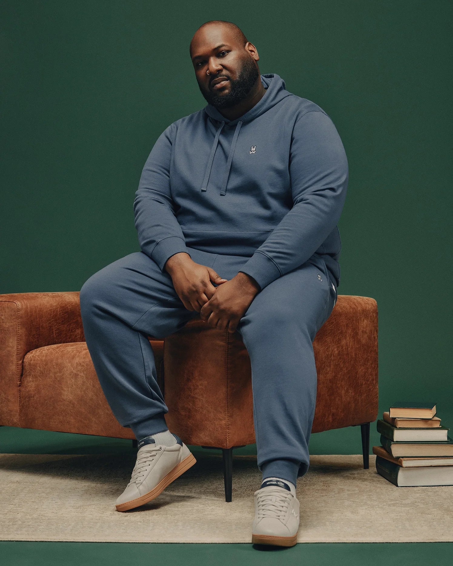 A man in a blue Psycho Bunny MENS BIG AND TALL LEVY ESSENTIAL FLEECE HOODIE (B9H859D200) sits on the arm of a brown chair against a green background, wearing gray sneakers, with stacks of books on the floor beside him.