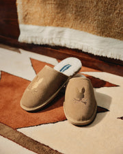 A cozy pair of MENS SUEDE SLIPPERS by Psycho Bunny, featuring a bunny and crossbones design, rests on a geometric-patterned rug in brown, white, and beige tones. A textured fabric in the background enhances their inviting charm.