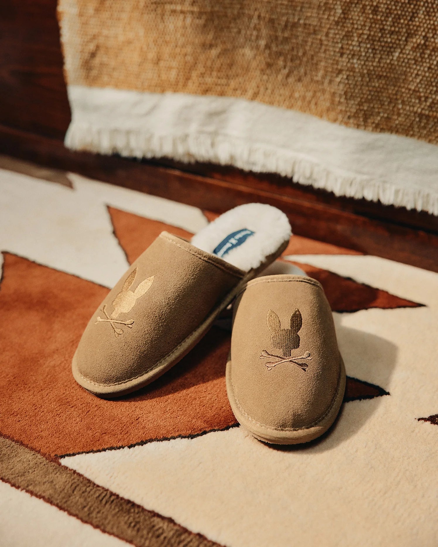 A cozy pair of MENS SUEDE SLIPPERS by Psycho Bunny, featuring a bunny and crossbones design, rests on a geometric-patterned rug in brown, white, and beige tones. A textured fabric in the background enhances their inviting charm.