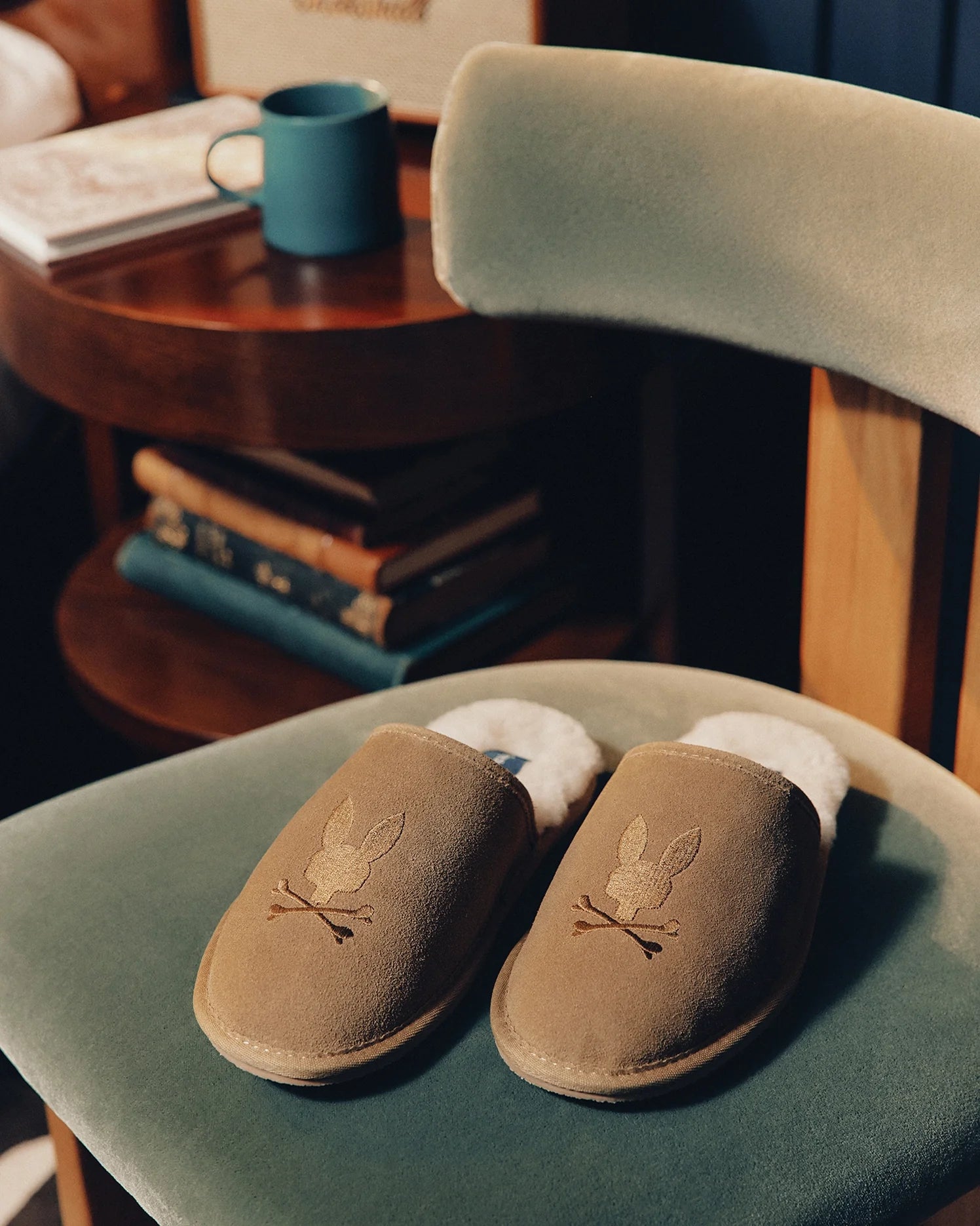 Psycho Bunny's MENS SUEDE SLIPPERS (B6A357E200) with a cozy rabbit design and genuine shearling lining rest on a green velvet chair. Nearby, a wooden side table holds a stack of books and a blue mug, creating an inviting atmosphere.