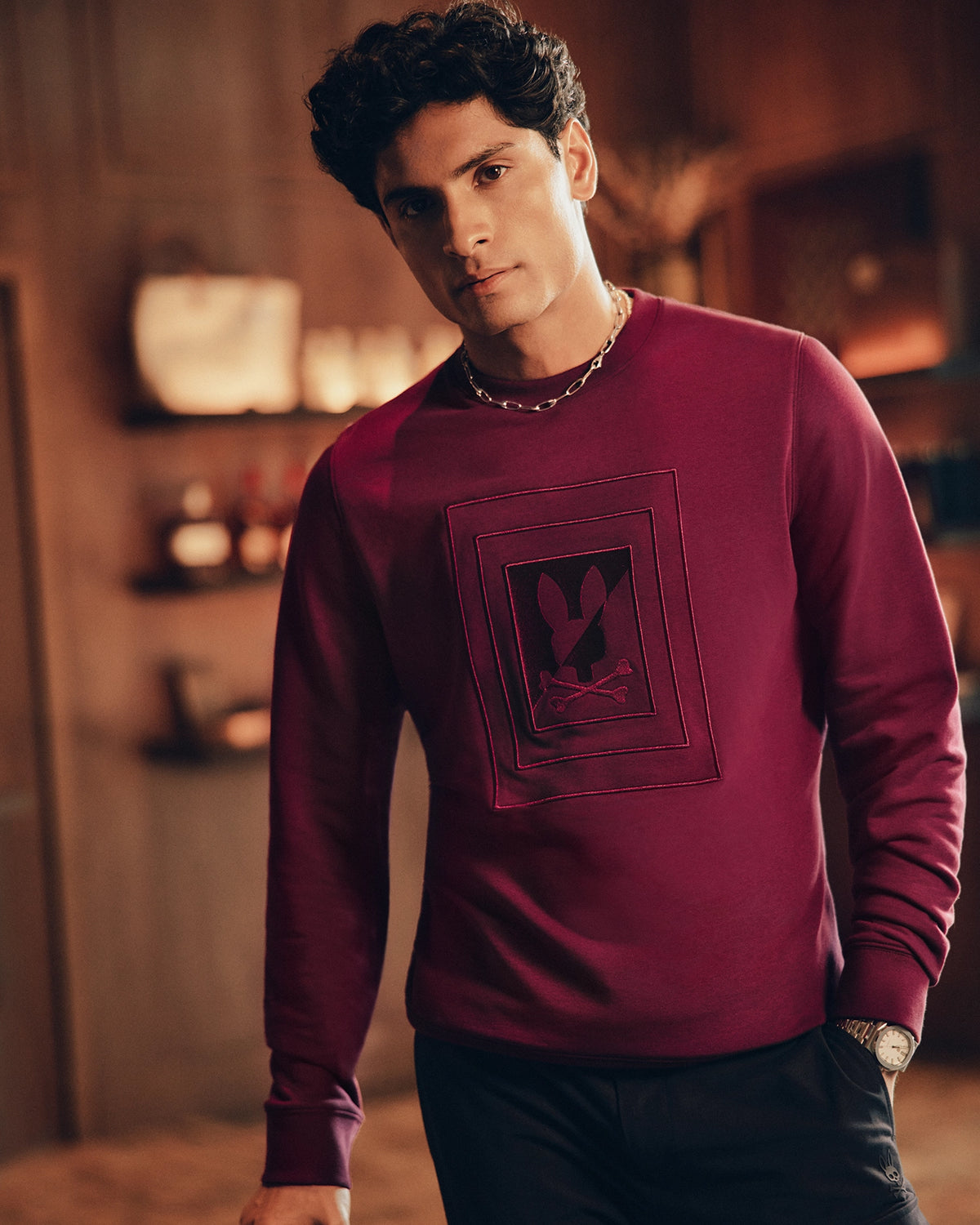A curly-haired person wears a maroon MENS OAK CITY CREW NECK - B6S274E200 by Psycho Bunny, featuring a black emblem, in a warmly decorated room with blurred shelves in the background.