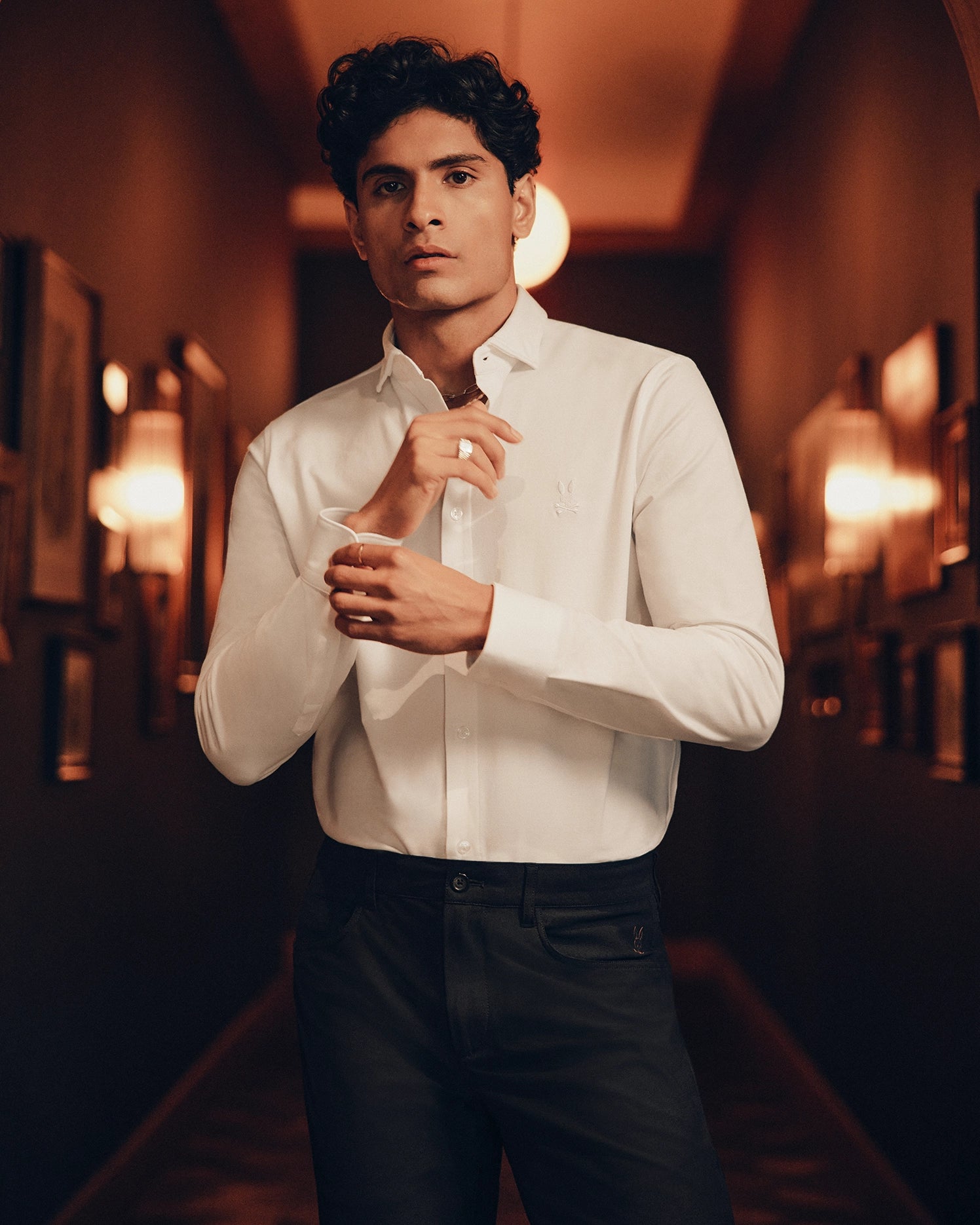 In a dimly lit hallway, an individual wearing the Psycho Bunny MENS ARTHUR JERSEY SHIRT - B6C748D200, made from stretch Pima cotton, is adjusting their shirt cuffs. They pair it with dark pants while framed pictures line the background walls.