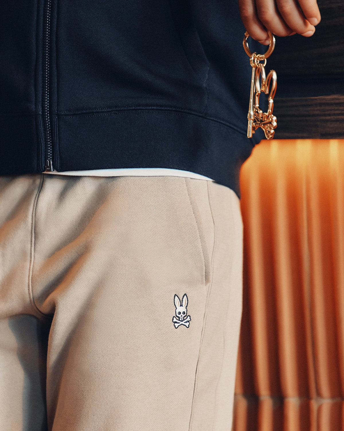 Close-up of someone holding gold keys with a decorative keychain. They're in cozy Psycho Bunny KIDS LEVY ESSENTIAL FLEECE SWEATPANT, featuring a small white rabbit and crossbones on the pocket, against warm-toned curtains and a dark jacket that enhance the snug vibe.