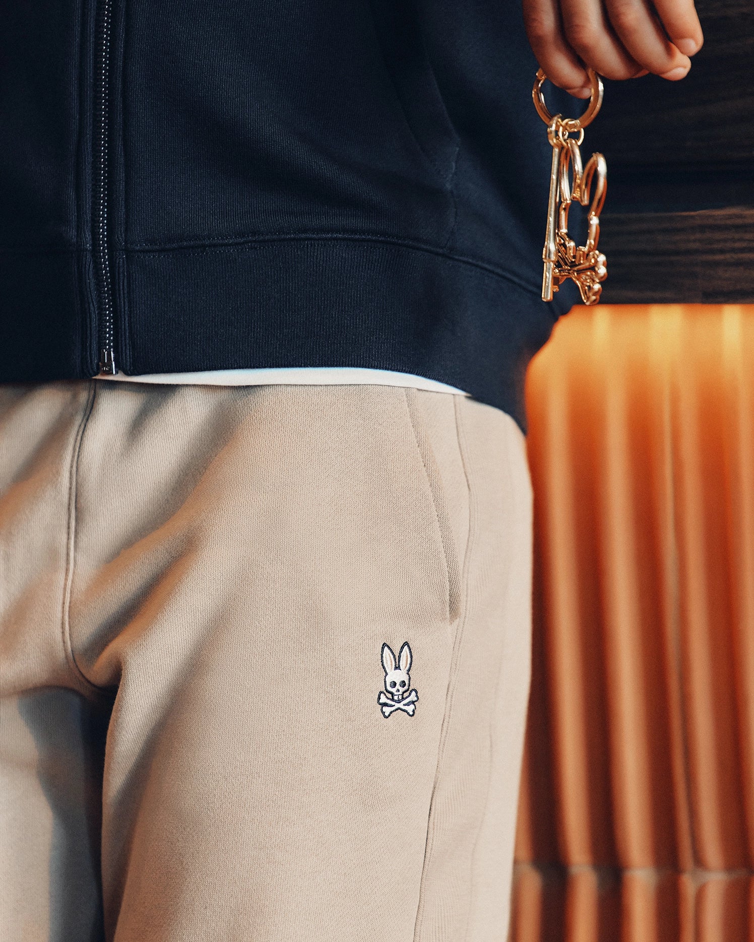 Close-up of someone holding gold keys with a decorative keychain. They're in cozy Psycho Bunny KIDS LEVY ESSENTIAL FLEECE SWEATPANT, featuring a small white rabbit and crossbones on the pocket, against warm-toned curtains and a dark jacket that enhance the snug vibe.