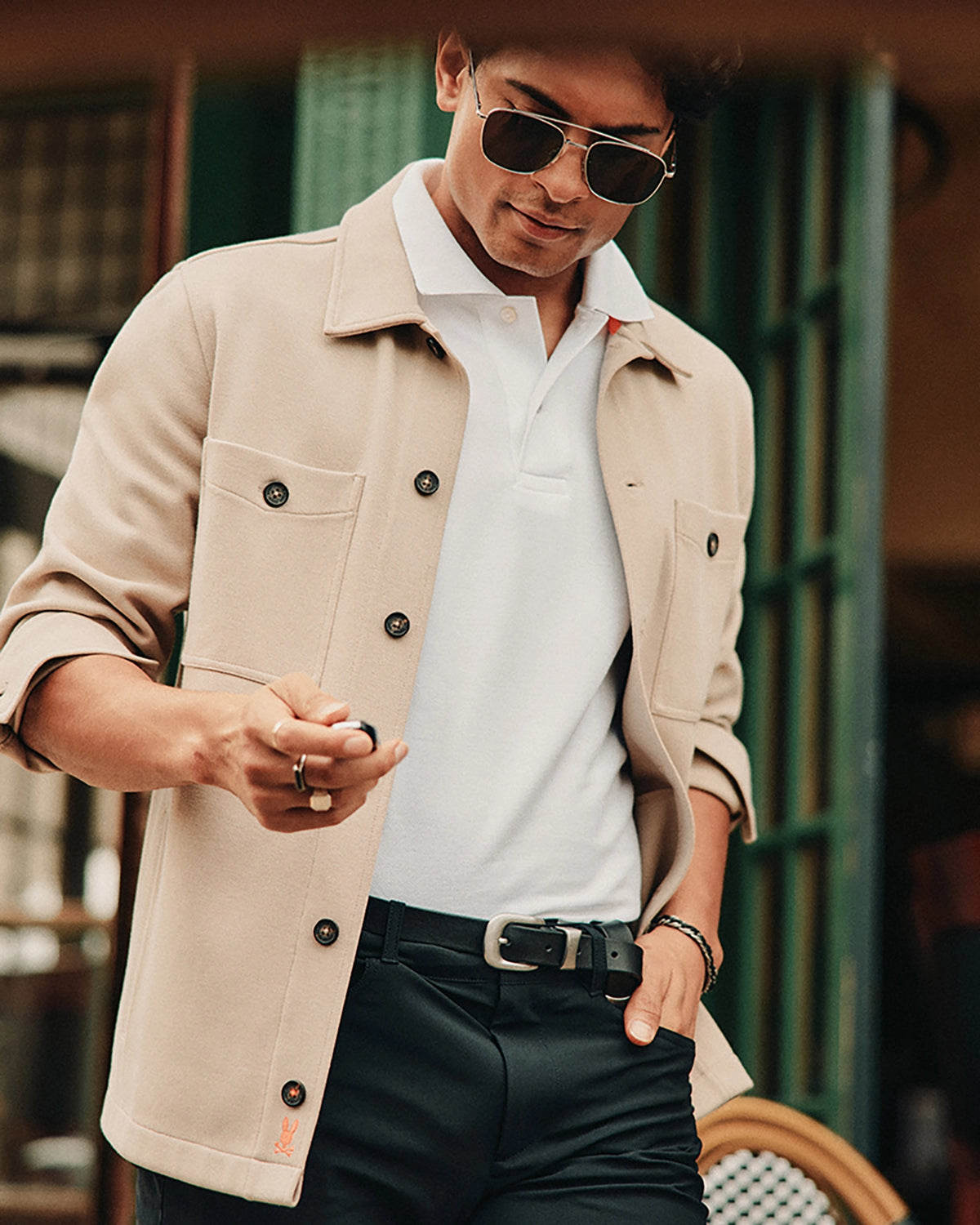 A person wearing sunglasses and a beige Psycho Bunny Men's Bedford Shirt Jacket (B6J290B200) over a white polo, featuring work-jacket style pockets, stands outdoors looking down. They're holding a smartphone against a contrasting green and white backdrop.