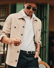 A person wearing sunglasses and a beige Psycho Bunny Men's Bedford Shirt Jacket (B6J290B200) over a white polo, featuring work-jacket style pockets, stands outdoors looking down. They're holding a smartphone against a contrasting green and white backdrop.