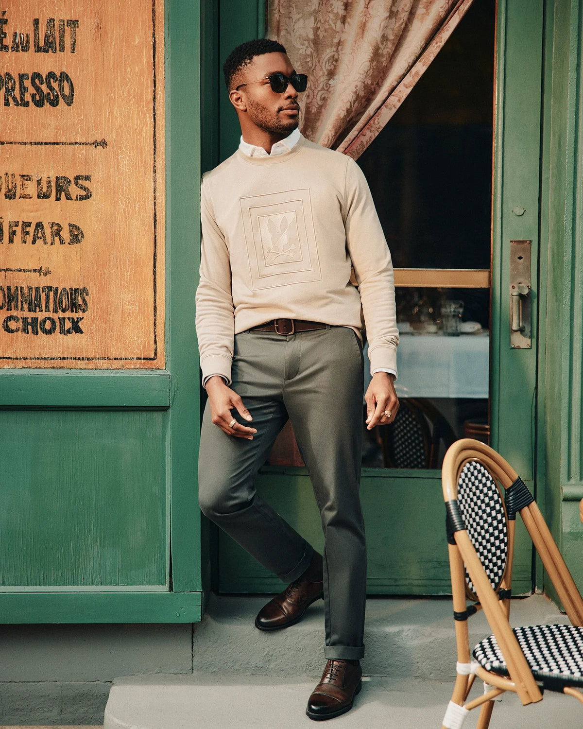 A man confidently stands outside a green-painted café, wearing sunglasses and the Psycho Bunny MENS OAK CITY CREW NECK sweater in beige. With one hand in his pocket, he leans slightly against the wall as a chair sits in the foreground.